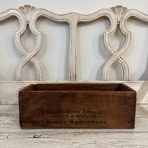 May include: A vintage wooden box with the text "Delco-Remy Lovejoy Hydraulic Shock Absorbers" printed on the front. The box is a dark brown color and appears to be aged. The background features a white, ornate chair back.