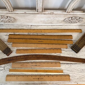 May include: A collection of vintage wooden measuring tools including several straight rulers with printed inch markings, two folding rulers with metal hinges, and one curved wooden drafting ruler, all featuring natural wood tones and dark text.
