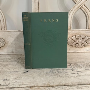 May include: A vintage hardcover book titled "FERNS" in a dark green color. The book has gold lettering and a decorative emblem. The spine reads "THE GARDEN LIBRARY" and "FERNS". The book is displayed on a distressed white wooden surface.