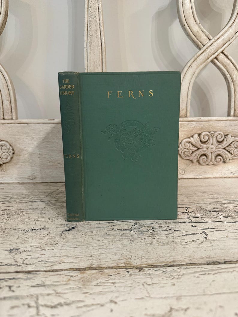 May include: A vintage hardcover book titled "FERNS" in a dark green color. The book has gold lettering and a decorative emblem. The spine reads "THE GARDEN LIBRARY" and "FERNS". The book is displayed on a distressed white wooden surface.