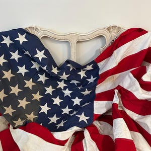 May include: An American flag with red and white stripes and a blue field with white stars is draped over a white, ornate chair back. The flag is vintage, with a slightly faded appearance.