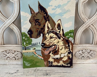 May include: A vintage paint-by-numbers artwork featuring two dogs. The dog on the left is a brown and black Great Dane, while the dog on the right is a brown and white German Shepherd. The dogs are painted against a blue sky with white clouds.