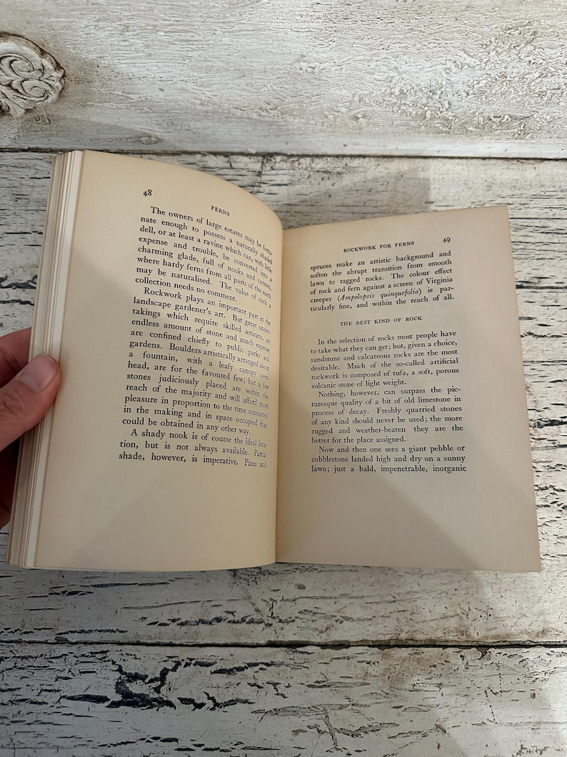 May include: Open book with pages displaying text about ferns and rockwork. The book is held open by a hand, revealing the text and the aged paper. The pages are yellowed with age, and the book is open on a wooden surface.