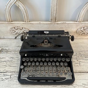 May include: A black Royal typewriter with silver keys. The typewriter has a classic design with a carriage return lever and a paper feed roller. The word "ROYAL" is printed on the top. The typewriter is on a white distressed surface.