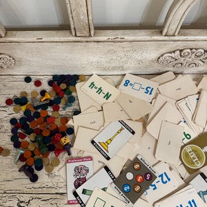 May include: A collection of vintage board game pieces and cards, including game cards with the words "Candlestick", "Professor Plum", and "Billiard Room". There are also colorful game tokens and dice scattered around.
