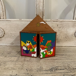 May include: A wooden house-shaped ornament with a star and a comet on the roof. The sides of the house are painted with a colorful scene of animals, houses, and a sun.