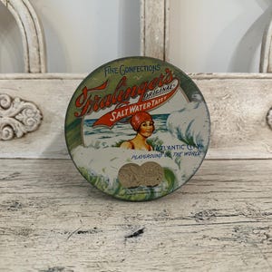 May include: Vintage round tin of Fralinger's Salt Water Taffy. The tin features a retro illustration of a woman in a red swim cap in the ocean. The text on the tin reads "Fine Confections" and "Atlantic City Playground of the World."
