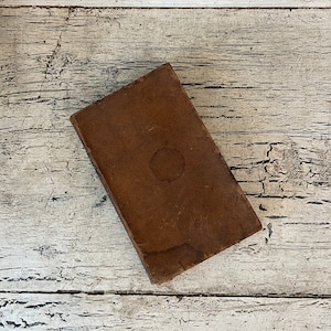 May include: A brown leather-bound book with a worn, aged appearance. The book is lying flat on a white wooden surface.