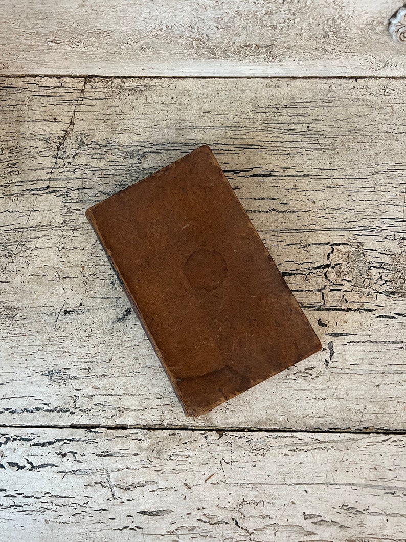May include: A brown leather-bound book with a worn, aged appearance. The book is lying flat on a white wooden surface.