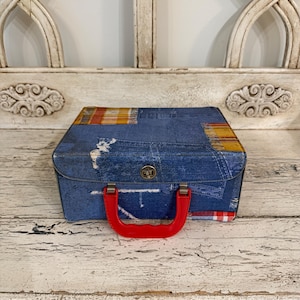 May include: A rectangular storage case featuring a blue denim-look exterior with printed patches of yellow and red plaid fabric. The case includes a bright red plastic handle, a metal latch closure on the front, and a structured, boxy design.