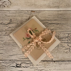 May include: A vintage book with a floral design on the cover is wrapped in a delicate pink lace ribbon. The book is sitting on a white wooden surface.
