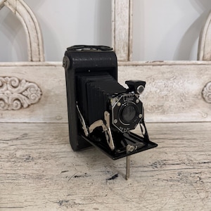 May include: A vintage folding camera with a black leather-covered body and a black accordion-style bellows. The front lens assembly is mounted on a metal plate with silver-toned mechanical supports and a small circular viewfinder on the top right.