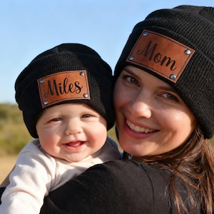 以下が含まれることがあります： 黒いニット帽に茶色の革パッチが付いています。片方の帽子には「Mom」、もう片方には「Miles」と書かれています。笑顔の女性と赤ちゃんが帽子をかぶっています。背景は野原です。