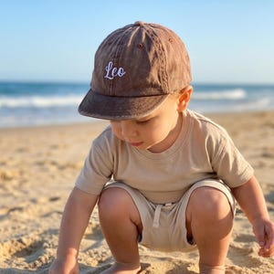 Könnte beinhalten: Braune Baseballkappe mit dem Namen "Leo" in Weiß gestickt. Ein Kind mit der Kappe ist an einem Sandstrand, mit dem Meer im Hintergrund. Das Kind trägt ein beiges T-Shirt und Shorts.