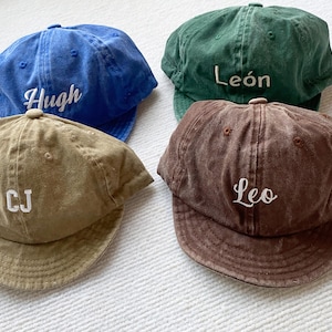 May include: Four baseball caps in shades of blue, green, tan, and brown. Each cap has a curved brim and embroidered text: "Hugh", "León", "CJ", and "Leo". The caps are made of a soft, textured fabric.