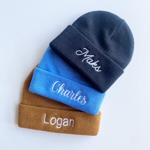May include: Three beanies in navy blue, light blue, and brown are stacked. Each beanie has a folded brim with white embroidered text: "Maks", "Charles", and "Logan". The beanies are made of a soft, knitted material.
