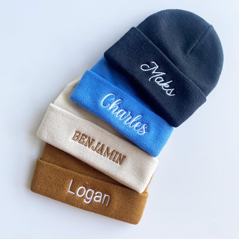 May include: A stack of four knit beanies in navy, blue, beige, and brown. Each beanie has a folded brim with embroidered text: "Maks", "Charles", "Benjamin", and "Logan" in white thread. The beanies are arranged diagonally.
