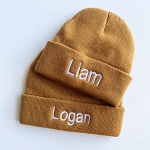May include: Two mustard-colored knit beanies are stacked on a white surface. The top beanie has a folded brim with the name "Liam" embroidered in white. The bottom beanie has the name "Logan" embroidered in white on its folded brim.