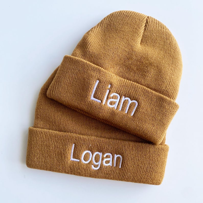 May include: Two mustard-colored knit beanies are stacked on a white surface. The top beanie has a folded brim with the name "Liam" embroidered in white. The bottom beanie has the name "Logan" embroidered in white on its folded brim.