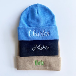 May include: Three stacked beanies in blue, navy, and tan. Each beanie features embroidered text: "Charles" in white, "Maks" in white, and "Tule" in green. The beanies are made of a soft, knitted material.