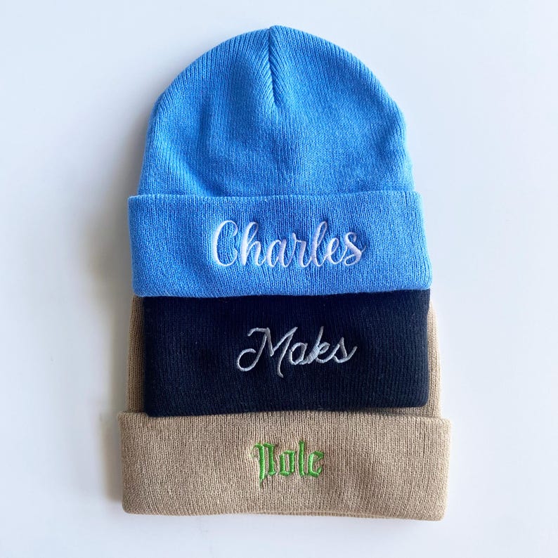 May include: Three stacked beanies in blue, navy, and tan. Each beanie features embroidered text: "Charles" in white, "Maks" in white, and "Tule" in green. The beanies are made of a soft, knitted material.