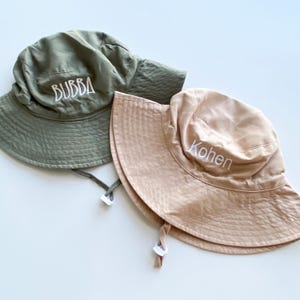 May include: Two sun hats. One is olive green with "BUBBA" embroidered in white. The other is tan with "Kohen" embroidered in white. Both hats have a wide brim and chin straps.
