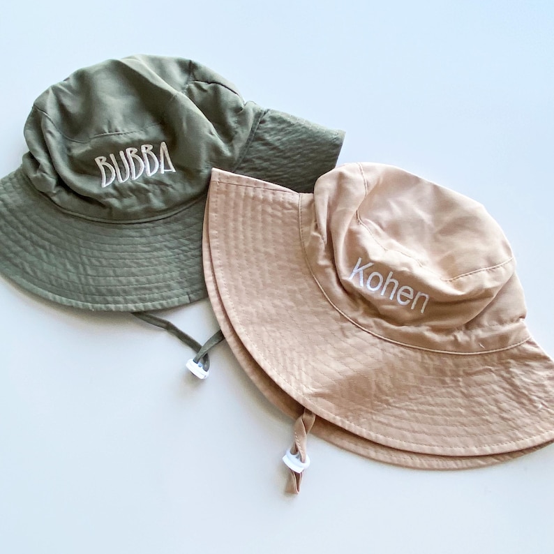 May include: Two sun hats. One is olive green with "BUBBA" embroidered in white. The other is tan with "Kohen" embroidered in white. Both hats have a wide brim and chin straps.