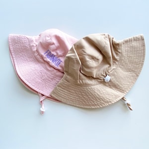 May include: Two bucket hats in the image. One is light pink with the name "FRANKIE" embroidered in purple. The other is a tan color. Both hats have a drawstring and a wide brim. The hats are made of a soft, textured fabric.