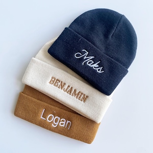 May include: Three knit beanies in navy, cream, and brown. Each beanie has an embroidered name: "Maks" in white, "Benjamin" in brown, and "Logan" in white. The beanies are stacked on a white surface.