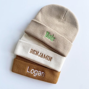 May include: A stack of three knit beanies in beige, cream, and brown. The top beanie has the name "Title" embroidered in green. The middle beanie has "BENJAMIN" embroidered in brown. The bottom beanie has "Logan" embroidered in white.