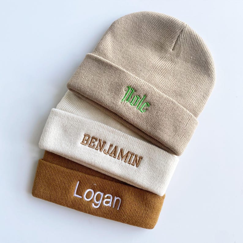 May include: A stack of three knit beanies in beige, cream, and brown. The top beanie has the name "Title" embroidered in green. The middle beanie has "BENJAMIN" embroidered in brown. The bottom beanie has "Logan" embroidered in white.