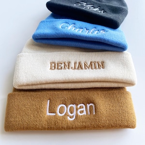 Peut inclure: Une pile de cinq bonnets en tricot de différentes couleurs : charbon, bleu clair, crème et moutarde. Chaque bonnet porte un nom brodé en couleur contrastante : "Maks", "Charles", "Benjamin" et "Logan".