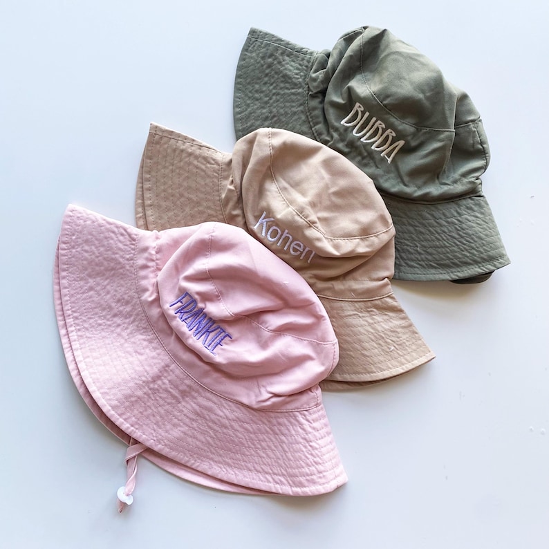 May include: Three bucket hats in shades of pink, tan, and olive green are displayed. Each hat has a personalized name embroidered on the front: "FRANKIE", "Kohen", and "BUBBA". The hats are made of a textured fabric and have a chin strap.