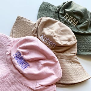 May include: Four bucket hats in shades of pink, tan, and green. Each hat has a different name embroidered in white or purple thread: "FRANKIE", "Kohen", and "BUBBA". The hats are made of a soft, textured fabric.