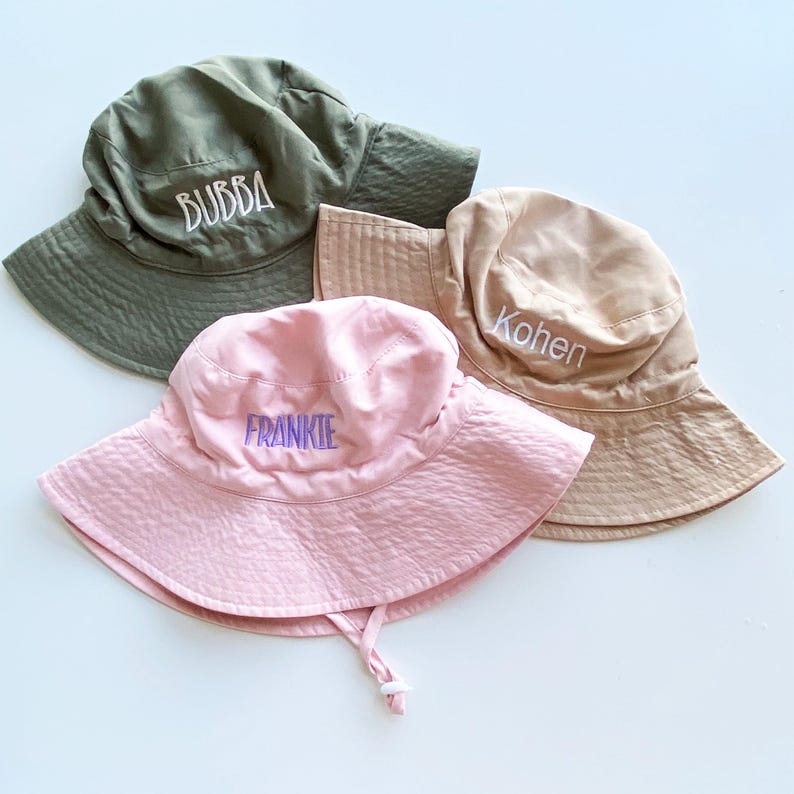 May include: Three personalized bucket hats in olive green, tan, and pink. Each hat features embroidered text: "BUBBA", "Kohen", and "FRANKIE". The hats have a wide brim and a chin strap.