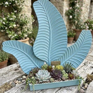May include: A light blue rectangular metal planter filled with various succulents, including green, purple, and silver rosettes. Three large, light blue, leaf-shaped metal decorative accents stand behind the plants. A small gold sign reads Succulent Garden.
