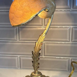 May include: A table lamp featuring a gold-toned base with a fluted, scalloped design and a stem shaped like a leaf. The shade is a translucent, peach-colored glass dome with a textured, scale-like pattern, accented by gold metal leaf details.