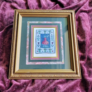 May include: A framed artwork featuring a central illustration of a figure in a pink dress under a crescent moon, surrounded by small green figures and floral motifs. The piece is encased in a gold-toned frame with a dark green and patterned mat.