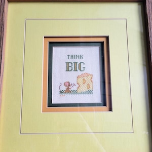 May include: A framed cross-stitch embroidery featuring the text "THINK BIG" in dark green and gold thread. Below the text, a small brown mouse stands next to a large yellow piece of Swiss cheese. The piece is set within a dark green, orange, and yellow mat.