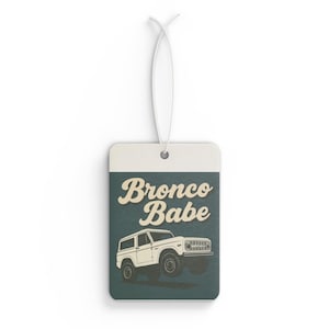 May include: A rectangular hanging air freshener with a white elastic string. The design features a dark teal background with the cream-colored text "Bronco Babe" above a line-art illustration of a white vintage SUV vehicle in profile.