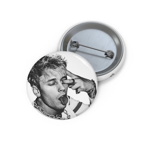 May include: Two white button pins. One pin features a black and white image of a person with their tongue out and a finger touching their eye. The other pin is plain.