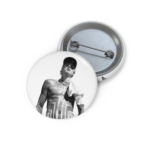 May include: Black and white button featuring a person wearing a baseball cap and holding a towel. The person has body art. A second button is visible in the background.