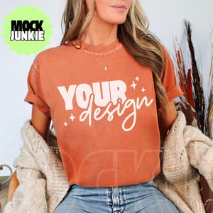 Peut inclure: Un t-shirt couleur rouille avec l'inscription "YOUR design" en blanc. Le t-shirt est porté avec un jean bleu et un cardigan crème. Une tasse avec un motif de feuilles est posée sur une surface en bois.