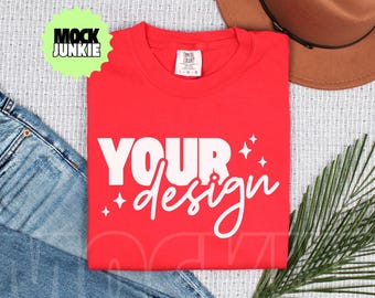 May include: A red t-shirt with the words "YOUR design" in white script and block letters, with star accents. The shirt is laid flat with a brown hat, blue jeans, and a sprig of greenery. The shirt has a tag that says "Comfort Colors".