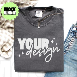 May include: A dark gray t-shirt with the words "YOUR design" in white script. A gold necklace with a small pendant rests on the shirt. The shirt is next to a pair of blue jeans and a plaid fabric.