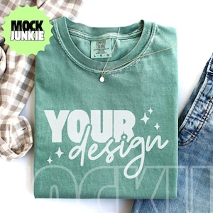 Puede incluir: Una camiseta verde salvia con las palabras "YOUR design" en escritura blanca y letras de bloque. Un collar de oro con un pequeño colgante descansa sobre la camisa. La camisa se combina con jeans azules y una camisa a cuadros.
