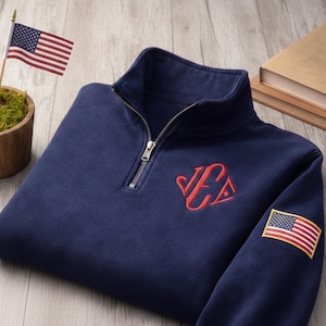 May include: Navy blue quarter-zip sweatshirt with a red embroidered monogram and an American flag patch on the sleeve. A small American flag is in the background. The sweatshirt has a silver zipper.