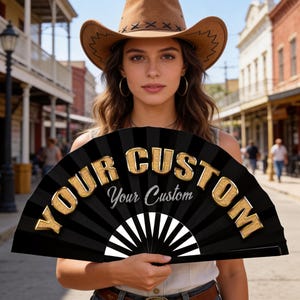 Op de afbeelding: Een zwarte opvouwbare handwaaier met de tekst "YOUR CUSTOM" in grote, gouden, driedimensionale letters en de woorden "Your Custom" in een kleiner, wit cursief lettertype. De waaier heeft een radiaal patroon van zwarte en witte strepen.