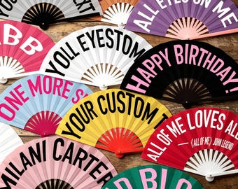 May include: A collection of handheld folding fans in various colours including red, yellow, pink, black, white, purple, and light blue. Each fan features bold black or white text with phrases like YOUR CUSTOM TEXT, ALL EYES ON ME, and HAPPY BIRTHDAY.