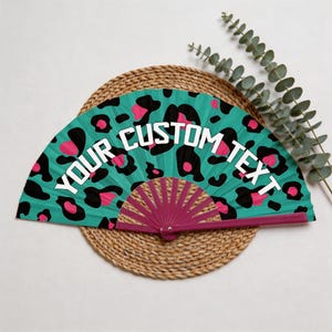 Op de afbeelding: Een turquoise handventilator met een luipaardprint in zwart en roze. De ventilator heeft de witte tekst "YOUR CUSTOM TEXT". De open ventilator ligt op een geweven mat, met groen in de achtergrond.